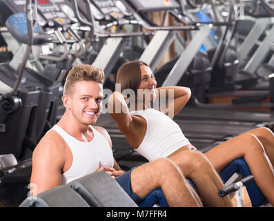 Homme bien formé coach aider cheerful fille pour faire l'exercice abs s'asseoir dans une salle de sport à l'intérieur de l'onduleur . L'accent sur l'homme Banque D'Images