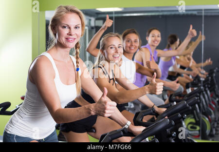 Smiling athletic girls giving Thumbs up lors de l'entraînement sur vélo stationnaire à gym Banque D'Images