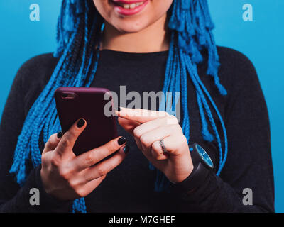 Adorable femme caucasienne hipster avec tresses africaines coiffure colorés contrôle news feed ou la messagerie via les réseaux sociaux, à l'aide d'une connexion wi-fi on mobile phone, smiling, posant sur le mur bleu Banque D'Images