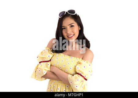 Fille asiatique en robe jaune avec des lunettes de soleil sur sa tête debout les bras croisés sur fond blanc. Banque D'Images