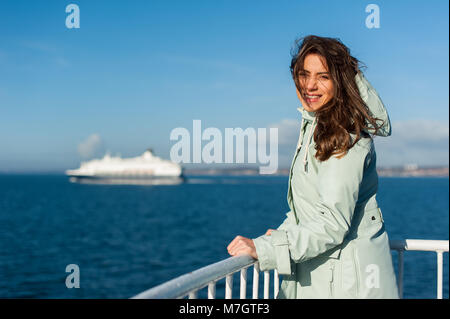 Jeune voyageur femme à la mer, de la voile d'un bateau avec gros bateau de croisière ou en ferry sur l'arrière-plan, portant une veste de pluie. Banque D'Images