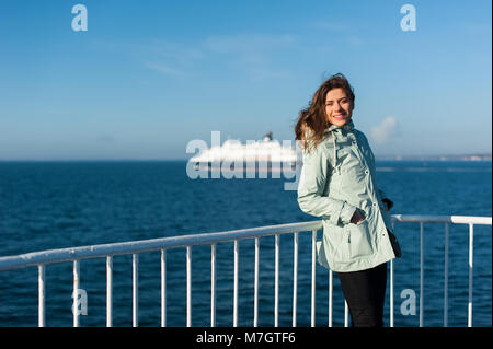 Jeune voyageur femme à la mer, de la voile d'un bateau avec gros bateau de croisière ou en ferry sur l'arrière-plan, portant une veste de pluie. Banque D'Images