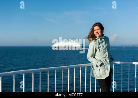 Jeune voyageur femme à la mer, de la voile d'un bateau avec gros bateau de croisière ou en ferry sur l'arrière-plan, portant une veste de pluie. Banque D'Images