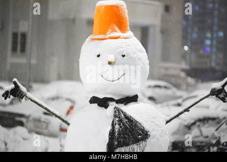 Bonhomme de neige sur un arrière-plan flou en gris avec feux bleus de la ville. Gros plan du bonhomme sourire avec bonnet, écharpe orange et carotte, nez à l'extérieur Banque D'Images