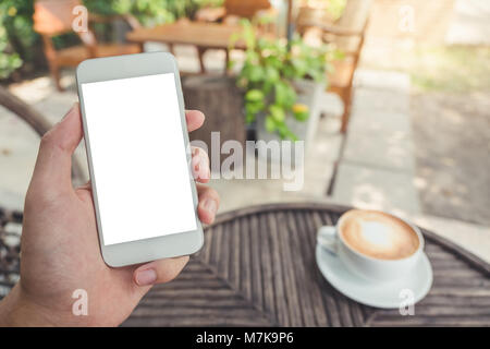 Immersive image de man's hand holding white téléphone mobile avec écran de bureau vierge dans un café avec l'arrière-plan flou outdoor Banque D'Images