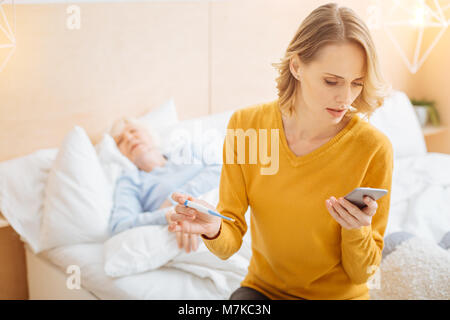 Jeune femme sérieuse d'essayer d'appeler un médecin en maintenant un thermomètre Banque D'Images