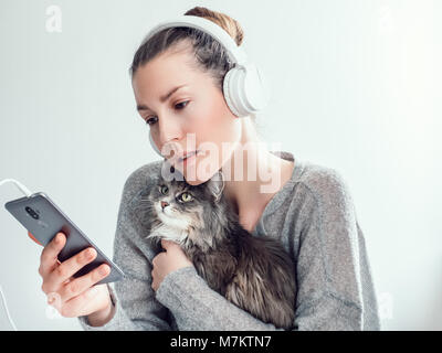 Jeune femme élégante, dans le casque et avec un téléphone mobile, l'étreindre doucement chaton sur un fond blanc. Les gens, des soins, de la technologie Banque D'Images