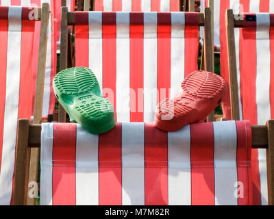 Enfant assis dans une chaise longue à rayures rouges et blanches avec leurs pieds, seulement les chaussures (une paire ou rouge et vert Crocs visibles) Banque D'Images