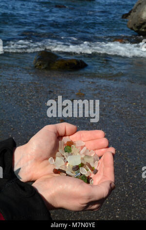 Une paire de mains femme tendre une poignée de verre de plage avec vagues se briser juste au-delà de son. Banque D'Images