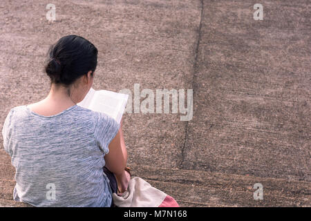 Une jeune femme se concentre de lire ici le dos dur livre tout en s'assit sur des mesures concrètes. Banque D'Images