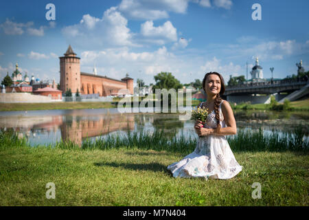 Jeune femme avec des fleurs dans l'habillement est assis sur l'herbe près de Kremlin et l'église dans une petite ville de la Russie. Belle fille sur la rive Banque D'Images