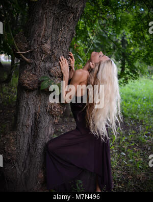 Jeune femme bien roulée béquilles sous le grand arbre et se détend dans le parc. Slim fille blonde avec de beaux cheveux dans l'habillement est de poser sur le fond de la nature Banque D'Images