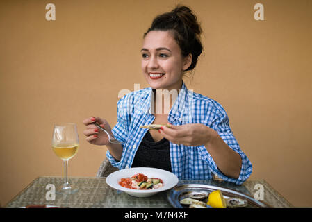 Jeune femme assise à table avec de la nourriture locale et verre de vin mousseux. Girl tient en mains oyster et fourche sur fond de mur jaune Banque D'Images