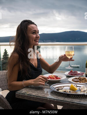 Jeune femme heureuse siège au balcon à table avec les huîtres, de la viande et de la salade et est titulaire d'un verre de vin mousseux. Fille élégante est de sourire et de rêver. Banque D'Images