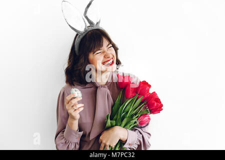 Belle fille élégante smiling avec oreilles de lapin et des tulipes roses holding easter egg sur fond blanc isolé. easter hunt concept. happy woman holdi Banque D'Images