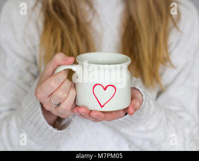 Une section d'une fille aux cheveux blonds tenant une tasse de café chaud avec un coeur d'amour sur tout en portant un chandail tricoté, chaleureuse dans un style confortable shot Banque D'Images