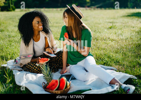 Deux jeunes fille multi-ethnique les amis sont heureux de parler et de manger la pastèque sur un pique-nique. La charmante enu africain l'un est maintenant le mobile. Banque D'Images