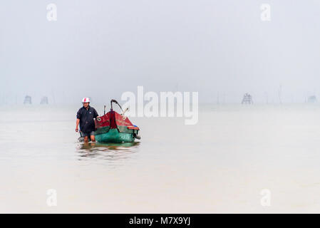 Un pêcheur vietnamien patauge dans les eaux peu profondes pour tirer son bateau de pêche traditionnel de la côte à la ferme ostréicole, Lap un lagon, Vietnam Banque D'Images