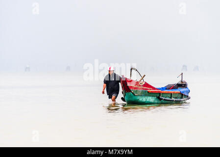 Un pêcheur vietnamien patauge dans les eaux peu profondes pour tirer son bateau de pêche traditionnel de la côte à la ferme ostréicole, Lap un lagon, Vietnam Banque D'Images