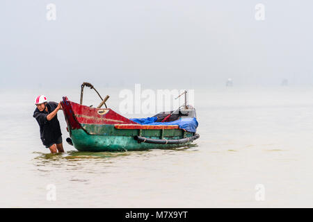 Un pêcheur vietnamien patauge dans les eaux peu profondes pour tirer son bateau de pêche traditionnel de la côte à la ferme ostréicole, Lap un lagon, Vietnam Banque D'Images