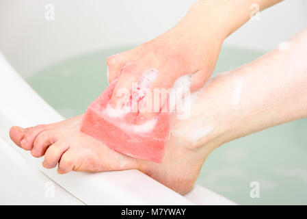 Caucasian woman lave sa jambe et pied dans une baignoire à l'aide d'une éponge de bain rose. Banque D'Images