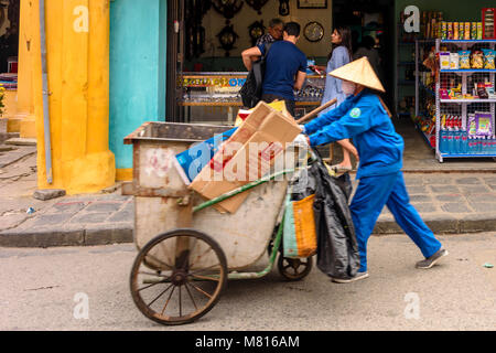 Une balayeuse portant un chapeau conique traditionnel bambou, pousse une brouette pour la collecte de déchets dans Hoi An, Vietnam Banque D'Images