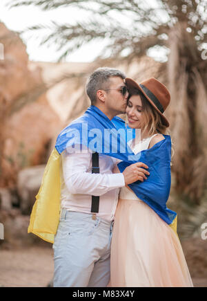 Jeune couple enveloppé dans un drapeau de l'Ukraine sont hugging in canyon contre fond de palmiers et de rochers. Libre. Banque D'Images