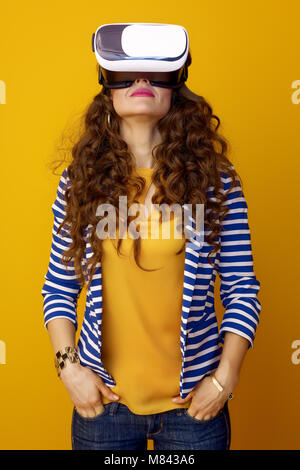 Femme à la mode avec de longs cheveux brun ondulé sur fond jaune portant des lunettes de réalité virtuelle Banque D'Images