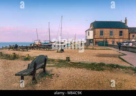 Whitstable, Kent, UK Beach avec des bancs en bois et de vieux bateaux en bois amarré sur la plage. Il y a un bâtiment en brique rouge et un sentier pour piétons. Il y Banque D'Images