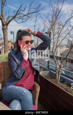 Belle jeune femme assise à l'extérieur et talking on mobile phone. Pour l'heure, profitant, de vie. Banque D'Images