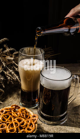Bouteille de bière mousseuse versée dans le verre de bière avec mug permanent avec du blé et panier de bretzels Banque D'Images