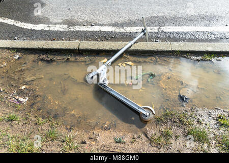 Un scooter de l'enfant est laissé à l'abandon dans une flaque d'eau boueuse où l'herbe point devrait être au bord d'une route. Banque D'Images