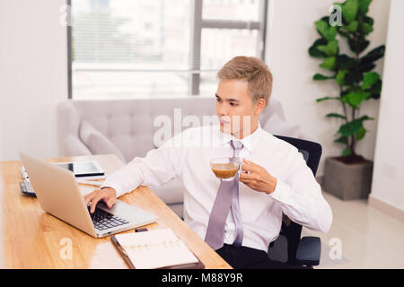Tourné à l'intérieur de l'entreprise créative avec bras tatoués, travailleur a barbe épaisse, est titulaire d'tasse de thé, l'air heureux en écran d'ordinateur portable, des messages avec frie Banque D'Images