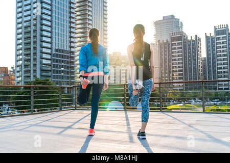 Vue arrière de deux jeunes femmes en bonne santé avec queue de l'étirement des jambes avant de courir pour rester en forme avec la lumière du soleil du matin ou du soir et vue sur la ville en t Banque D'Images