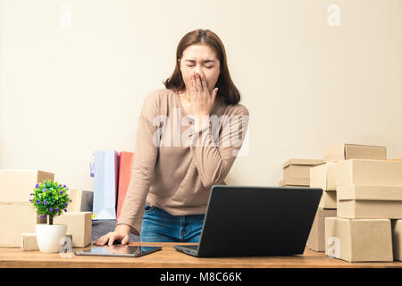 Young Asian woman yawning en ligne d'affaires tout en travaillant sur son ordinateur dans son bureau à domicile, entouré par des boîtes de ses produits, une bonne idée pour onli Banque D'Images