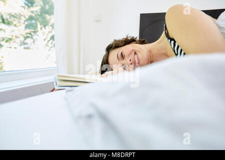 Mid adult woman, détente sur lit, holding book, smiling Banque D'Images