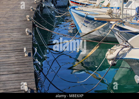 Différents bateaux à moteur amarrés sur une jetée en bois marron avec des cordes à des bitts de rivage sur l'eau de mer reflétant la lumière du soleil en été Banque D'Images