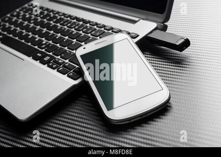 Téléphone blanc à blanc avec reflet vert s'appuyant sur une entreprise à côté d'un clavier d'un ordinateur portable branché en stockage USB Flash Drive, tous les au-dessus d'une couche de carbone Banque D'Images