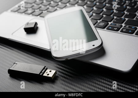 Smartphone blanc avec reflet vert couché sur le clavier de l'ordinateur portable d'affaires à côté d'un périphérique de stockage USB Flash Drive, tous les au-dessus d'une couche de carbone Banque D'Images