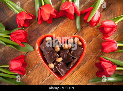 Bol en forme de coeur rouge avec des morceaux de chocolat et de tulipes rouges se trouvant dans un cercle autour de lui, sur le vieux fond de bois rustique de diagonale. Banque D'Images