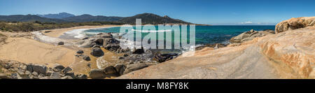 Vue panoramique d'ondes lave sur des rochers et du sable à la plage d'Algajola en Balagne Corse sous un ciel bleu profond avec des montagnes enneigées Banque D'Images