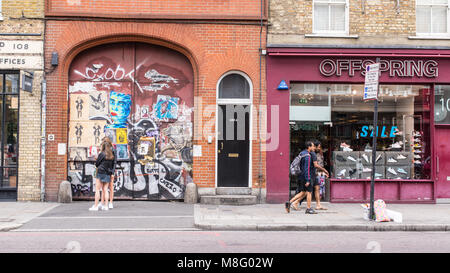 Personnes marchant devant une vieille porte couverts en autocollants et street art peintures murales à côté de progéniture en magasin Commercial Street, Spitalfields, Shoreditc Banque D'Images