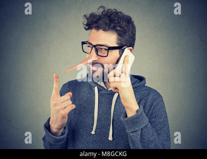 Jeune homme rusé avec long nez talking on mobile phone isolé sur fond de mur gris. Menteur concept. L'émotion des sentiments Banque D'Images