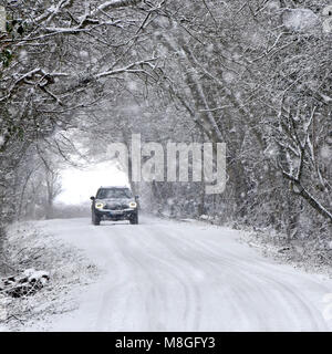 Phares de voiture allumés conduite à travers la neige tombante sur la route de campagne glacée neige couvert tunnel d'arbre dans la scène boisée arbres enneigés hiver météo Angleterre Royaume-Uni Banque D'Images