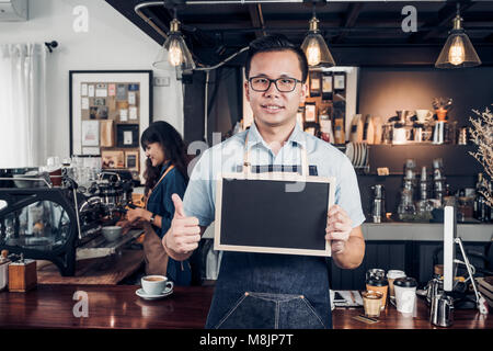 Asian male barista de porter un tablier jean Thumbs up at blank blackboard menu café au comptoir bar avec smile face,cafe service concept,propriétaire de l'entreprise start Banque D'Images