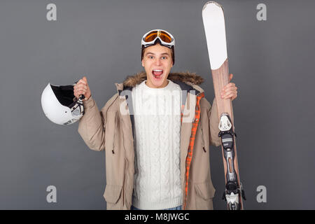 Jeune mec des vêtements d'hiver studio isolé sur mur gris holding skis helmet et à la surprise excitée de l'appareil photo Banque D'Images
