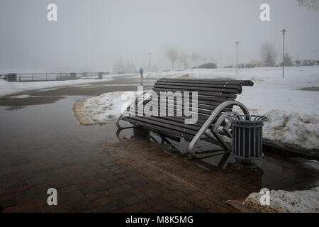Silhouette floue d'un homme au loin caché par le brouillard. Banc de parc près du sentier avec les flaques de neige fondue dans un brouillard épais. Début du printemps landsca Banque D'Images