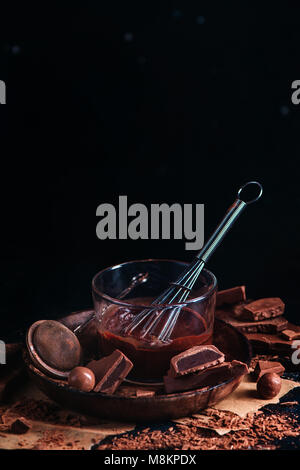 Vitrage de chocolat fondu dans une tasse en verre avec un fouet. Le chocolat noir et le chocolat au lait avec morceaux de cannelle et autres épices sur un fond noir. Banque D'Images