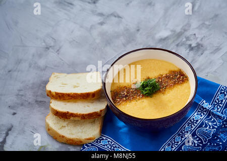 Bol en porcelaine violet de soupe de potiron crémeuse garnie d'aneth, servi avec des tranches de pain grillé sur bure sur serviette fond texturé blanc, Banque D'Images