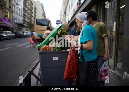 Femme noire handicapée avec béquille et d'autres personnes regardent à travers les déchets alimentaires dans la poubelle, supermarché extérieur, rue de Clignancourt, Paris, France Banque D'Images